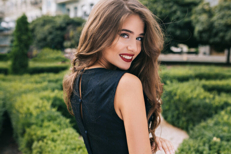 Closeup portrait from back of pretty girl with attractive makeup and red lips on street on green yard background. She wears black dress, has long curly hair